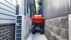 Slanetrac HT1000 mini dumper driving beside a house. It is passing through a narrow space carrying a large load of dirt. An operator stands behind it.
